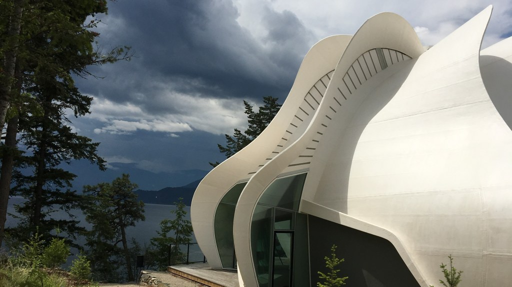A curved white building against a stormy backdrop of mountains and a lake.